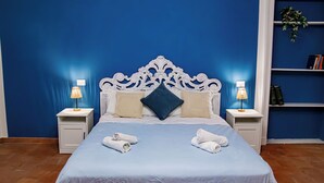 A neatly made bed with two pillows, a blue bedspread, and two rolled towels on the side. There are two bedside tables with lamps, and a bookshelf with books and a plant. - Cozy House with Lake View • Private Terrace & Self Check-in 24h (Viverone)