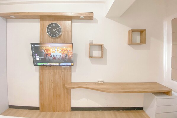 A modern room with a wooden reception desk, a mounted TV displaying a program, a clock, and a small wooden shelf.