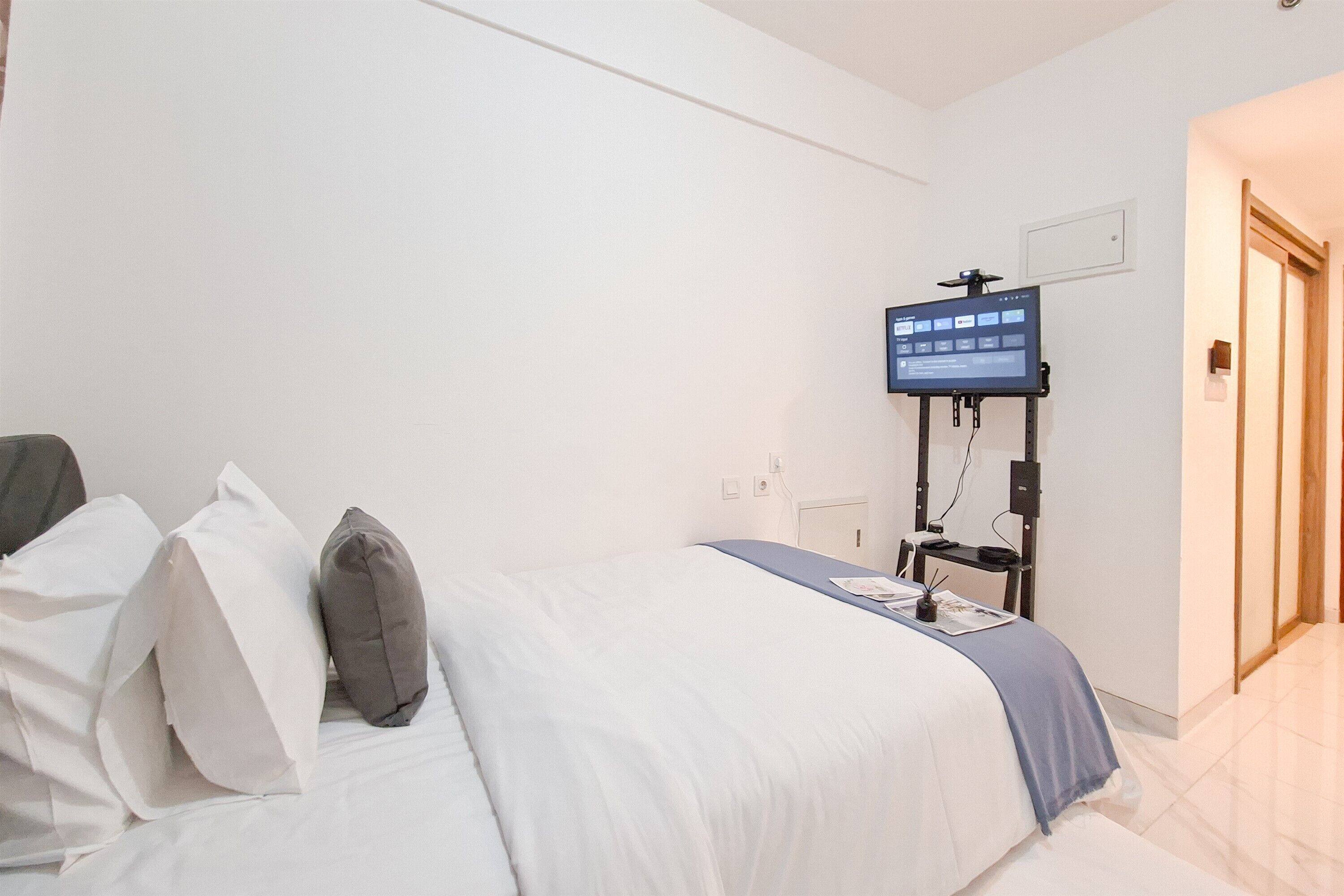 A hotel room with a bed, pillows, a TV mounted on the wall, and a door leading to another room.
