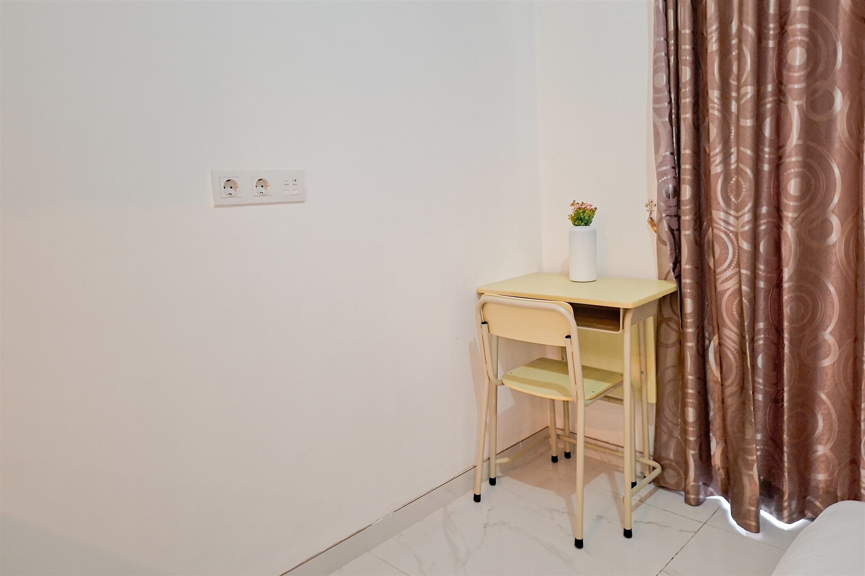 A small, single-bed room with a desk and chair, a power outlet on the wall, and a curtain on the window.