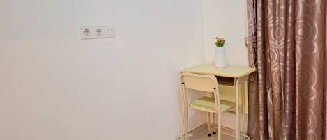A small, single-bed room with a desk and chair, a power outlet on the wall, and a curtain on the window.