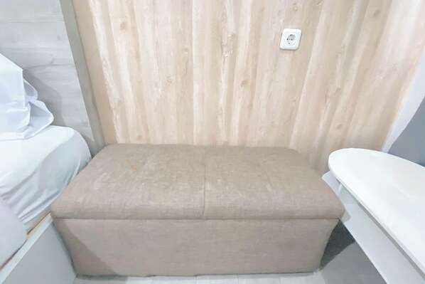 Un petit banc beige, recouvert de tissu, se trouve dans une pièce dont un mur est lambrissé et où un matelas blanc est posé sur la gauche.