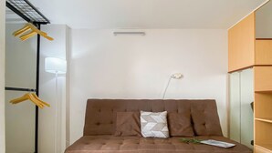 Ein braunes Schlafsofa mit einem weißen Kissen und einer Zeitschrift darauf, eine Stehlampe und ein Kleiderschrank mit Spiegel.