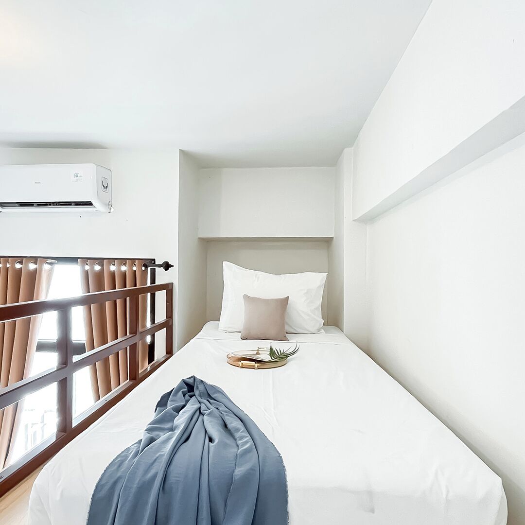 A small, white, single-bed room with a wooden headboard, a bedside table with a plant, and a window with curtains.