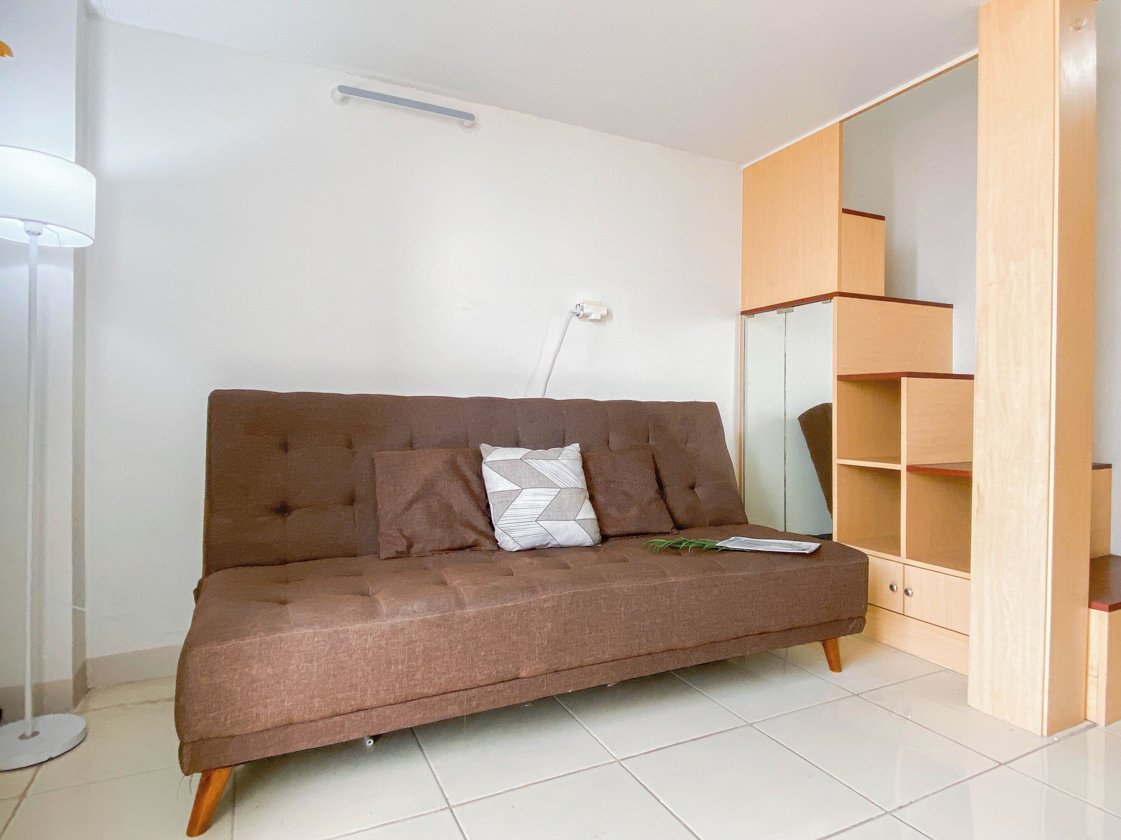 A brown sofa with cushions, a floor lamp, and a wooden shelving unit in a room with white walls and tiled flooring.