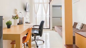 Ein kleiner, hell erleuchteter Raum mit Schreibtisch, Stuhl und einer Couch. An der Wand ist ein Fernseher, eine Topfpflanze und ein Fenster mit Vorhängen.