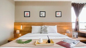 Ein Hotelzimmer mit einem hölzernen Kopfteil, weißer Bettwäsche, drei gerahmten Blumenbildern an der Wand, Nachttischlampen, einer Ablage mit einer Flasche und einer Fernbedienung sowie einer Zeitschrift auf dem Bett.