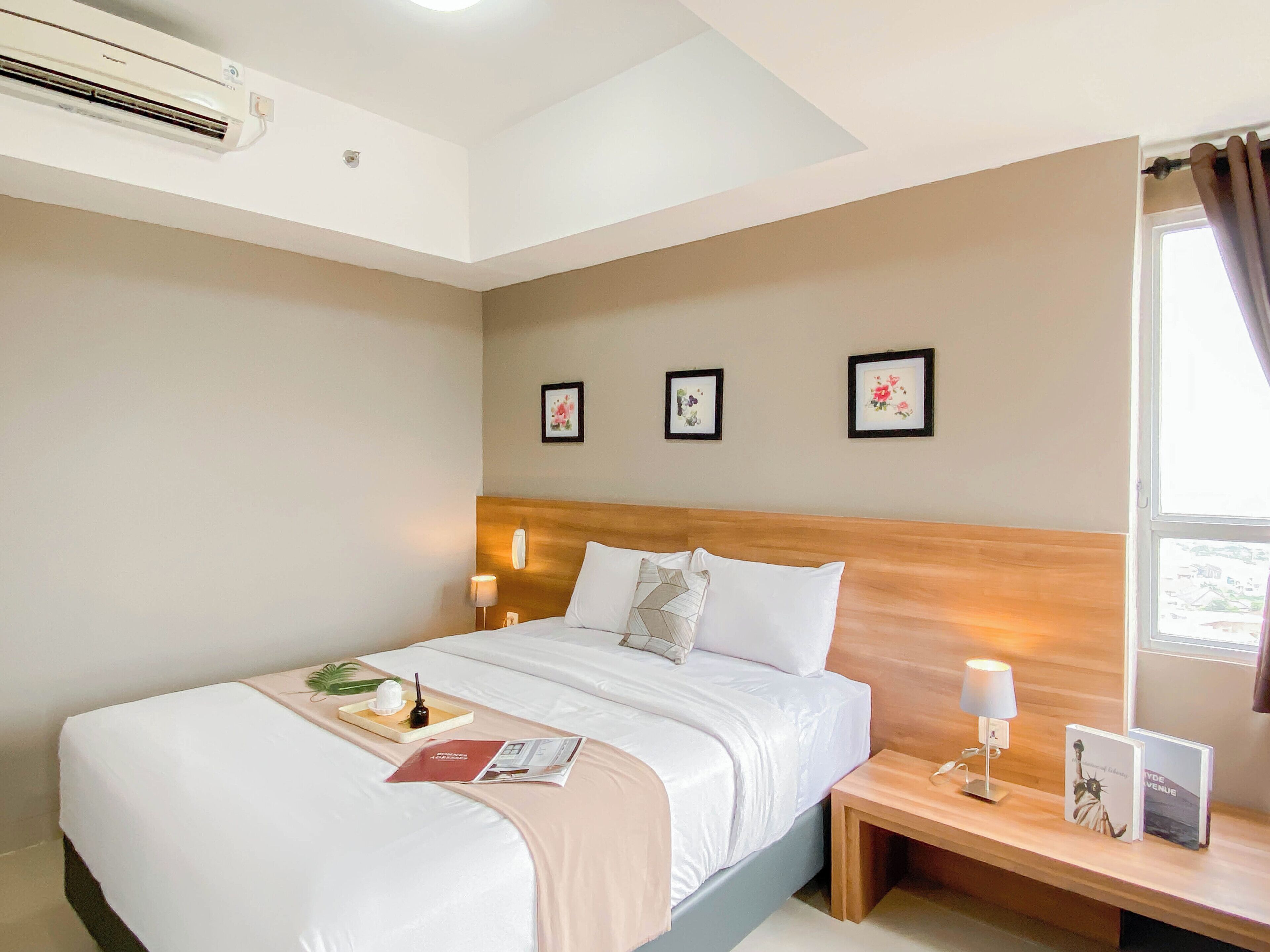 A hotel room with a bed, bedside tables, a nightstand, a wall-mounted air conditioner, and framed pictures on the wall.