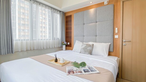 Une chambre d’hôtel avec un grand lit, une tête de lit en bois, une fenêtre avec des rideaux légers et une porte.