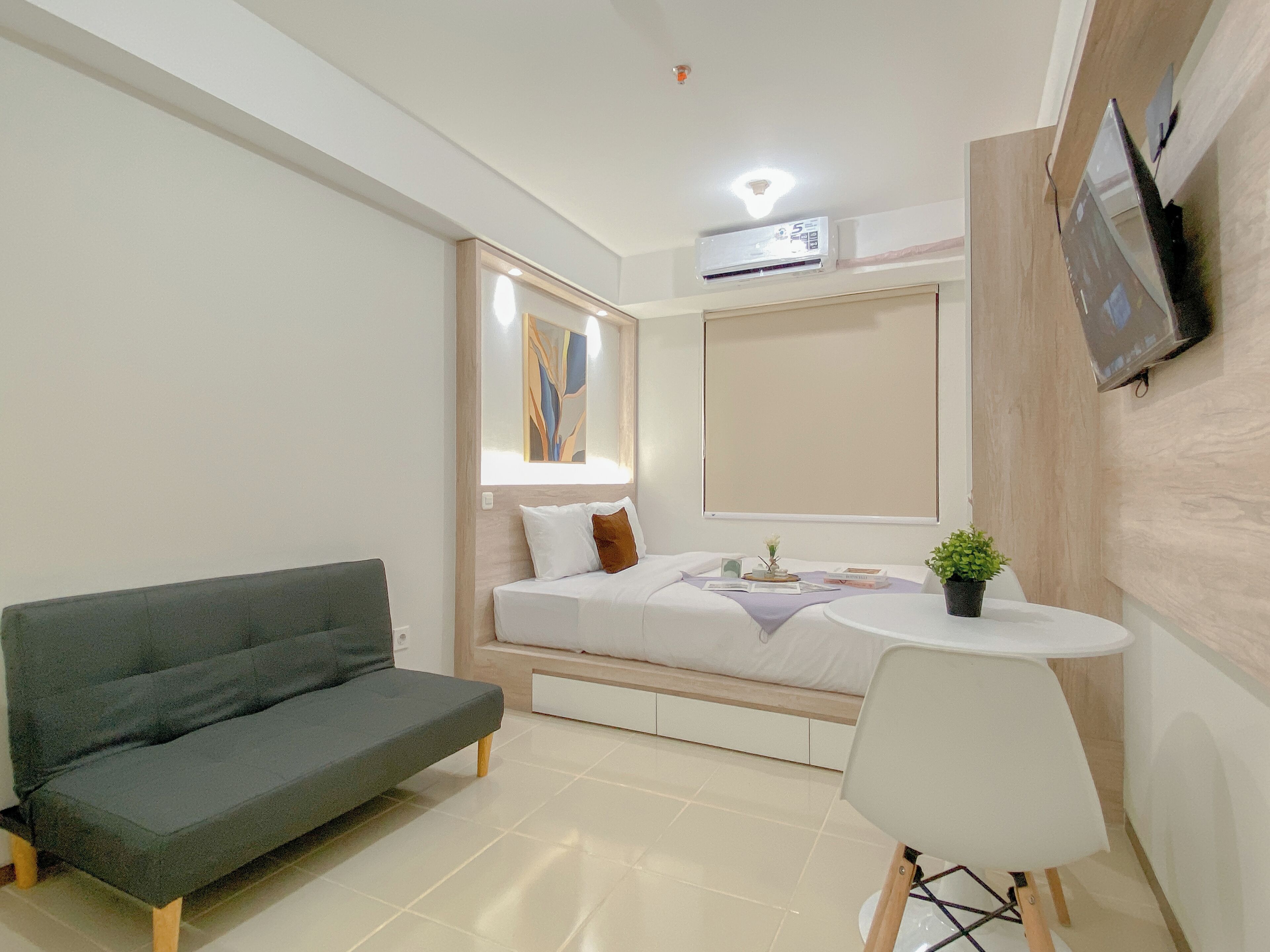 Ein modernes Hotelzimmer mit grauem Sofa, einem kleinen weißen Tisch, einem Bett mit weißem und braunem Kissen, einem wandmontierten Fernseher und einer Klimaanlage.