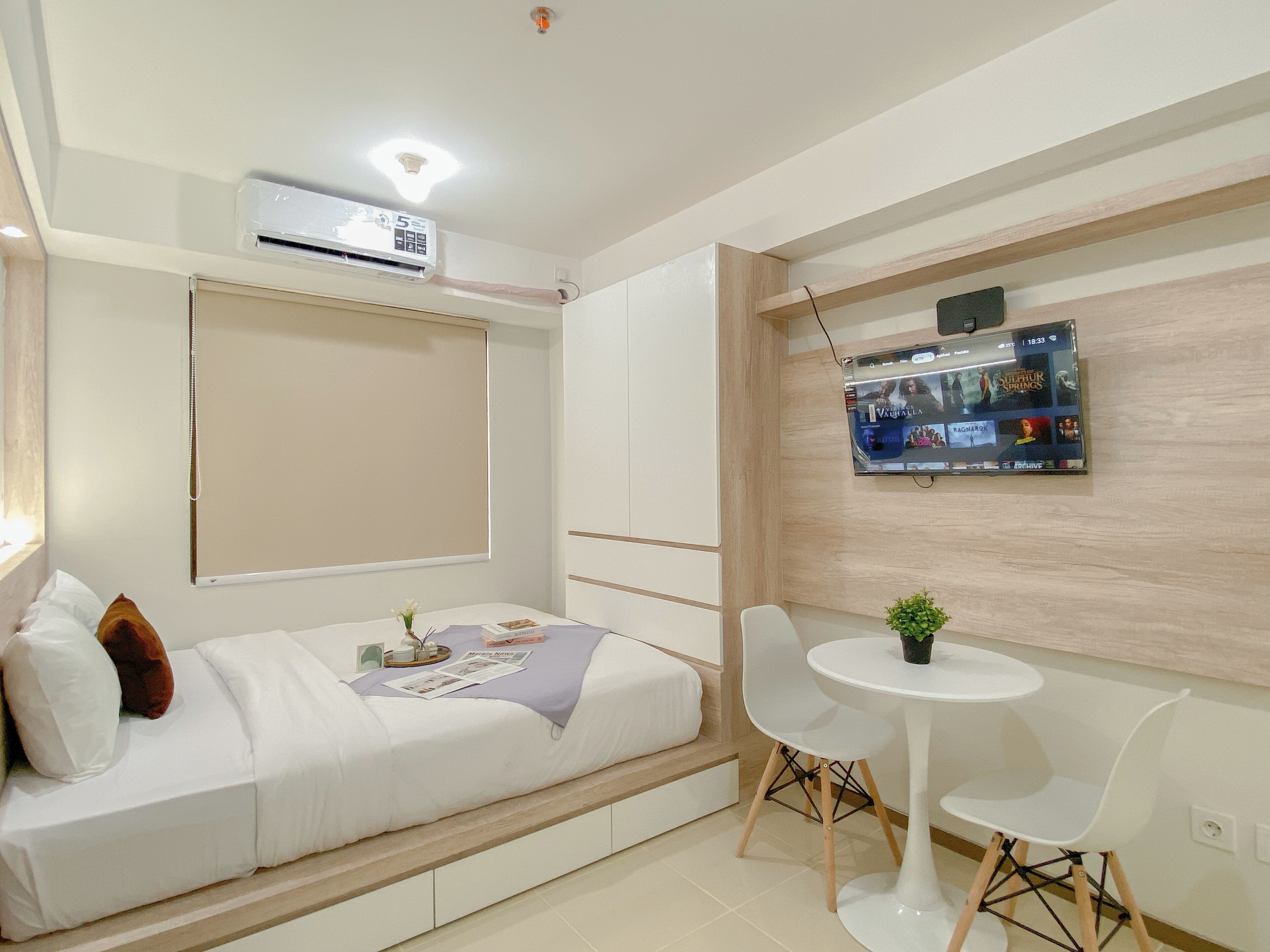 Ein modernes Schlafzimmer mit einem Bett, einem kleinen Tisch mit Stühlen, einem an der Wand befestigten Fernseher und einer Klimaanlage.