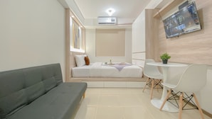 A modern hotel room with a grey sofa, a bed with white bedding, a small table with a plant, and a TV mounted on the wall. - Best Deal And Homey Studio At Meikarta Apartment (Cikarang)