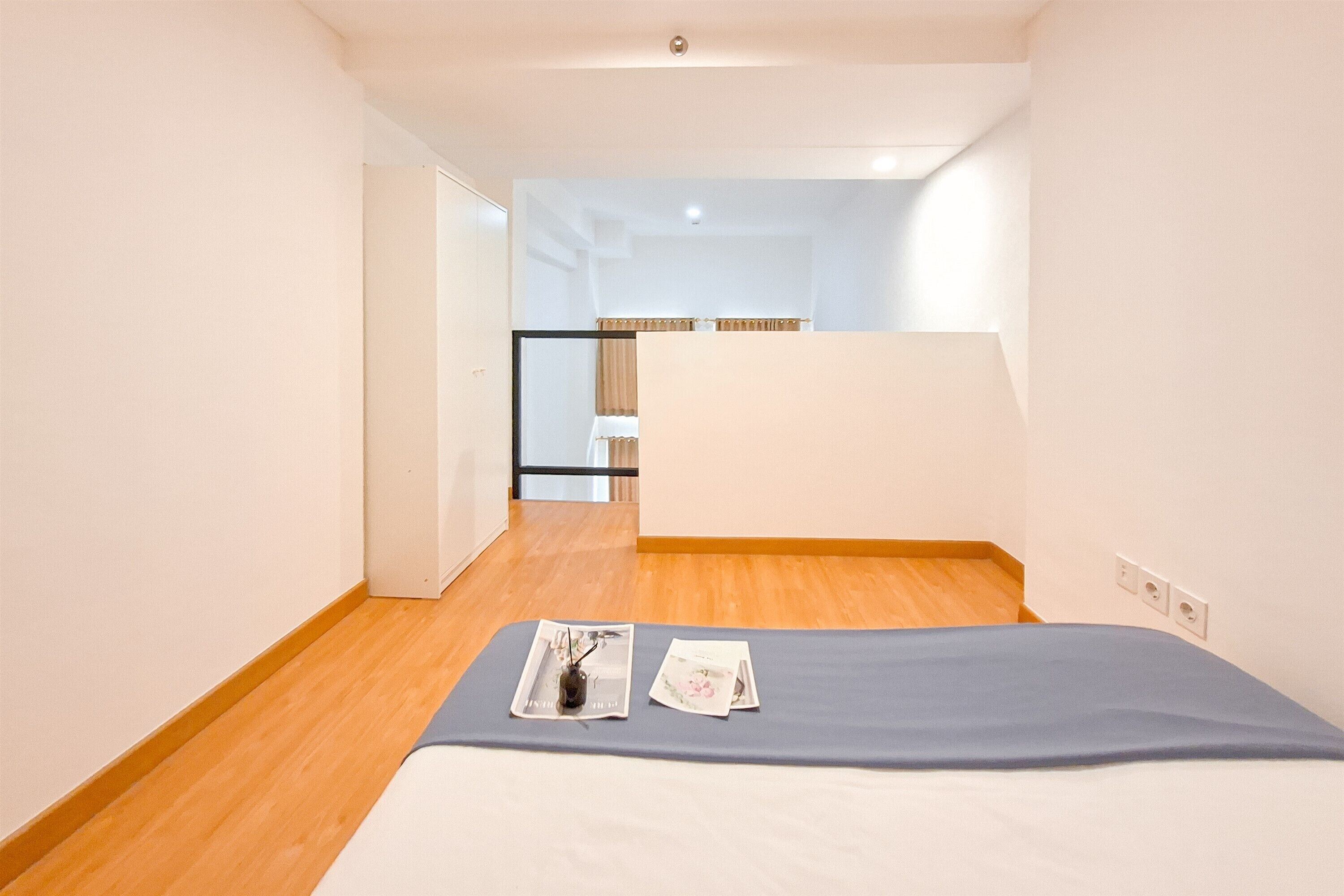 Una habitación de hotel con una cama, una mesita con una revista y un florero, y vistas al pasillo y otra habitación.
