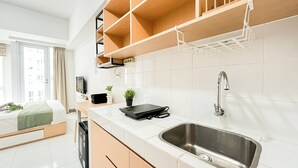 A modern kitchen with white tiles, a stainless steel sink, wooden shelves, a black countertop, and a window with curtains. - Good And Enjoy Living Studio At Tokyo Riverside Pik 2 Apartment (Teluknaga)
