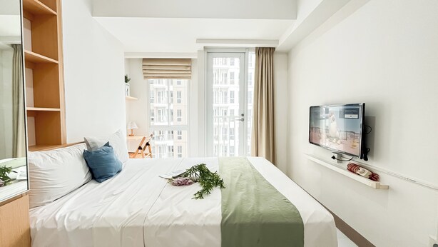 A hotel room with a bed, a TV mounted on the wall, and a view of the city through the window. - Good And Enjoy Living Studio At Tokyo Riverside Pik 2 Apartment (Teluknaga)