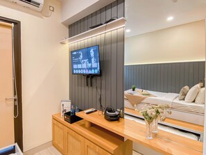 Une chambre d’hôtel moderne dotée d’un grand téléviseur à écran plat fixé au mur, d’un bureau en bois avec une machine à café et d’un lit avec une tête de lit grise.