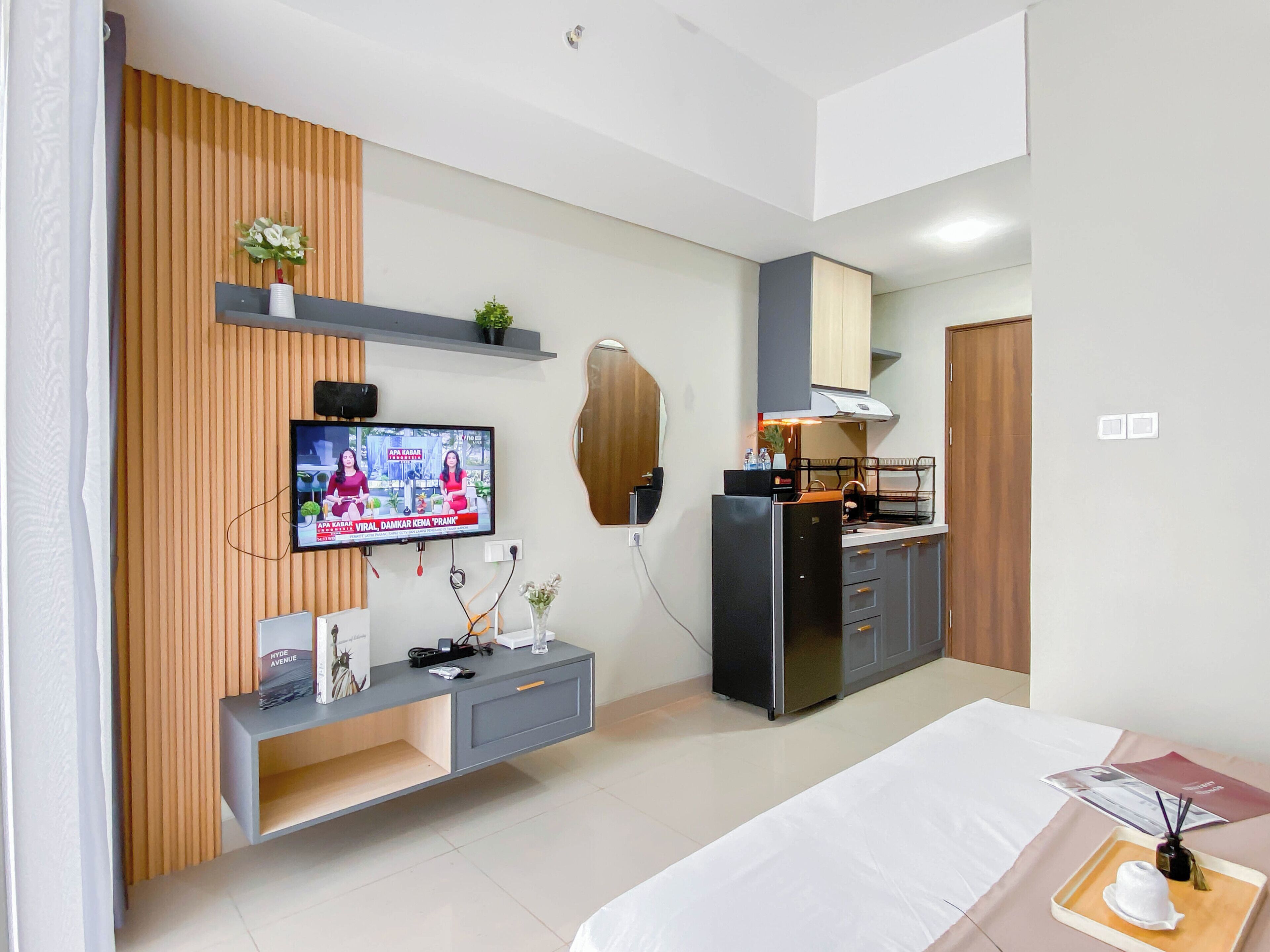 A modern hotel room with a flat-screen TV mounted on a wooden panel wall, a kitchenette with a refrigerator and microwave, a bed with a white bedspread, and a small table with a tray.