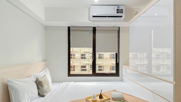 A hotel room with a bed, a window with a view of buildings, an air conditioning unit, and a bedside table with a tray. - Neat Studio Room Apartment Meikarta (Cikarang)