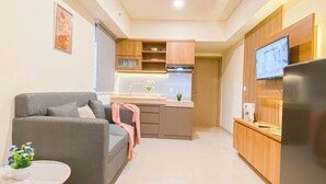 A modern living room with a grey sofa, a small round table with a plant, and a kitchen area with wooden cabinets and a TV mounted on the wall. - Comfy Apartment 2Br At Meikarta (Cikarang)