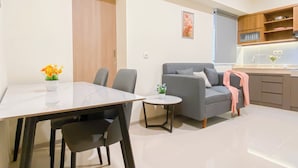 A modern living room with a dining table, grey sofa, and a small kitchen area with wooden cabinets and a sink. - Comfy Apartment 2Br At Meikarta (Cikarang)
