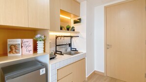 A modern kitchen with wooden cabinets, a stainless steel sink, and a built-in oven. There is a small refrigerator and a shelf with books and a vase of flowers. - Restful And Tidy Studio At Pollux Chadstone Apartment (Cikarang)