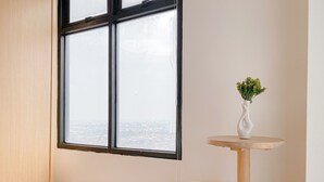 A room with a large window, a small round wooden table, and a white stool. - Restful And Tidy Studio At Pollux Chadstone Apartment (Cikarang)