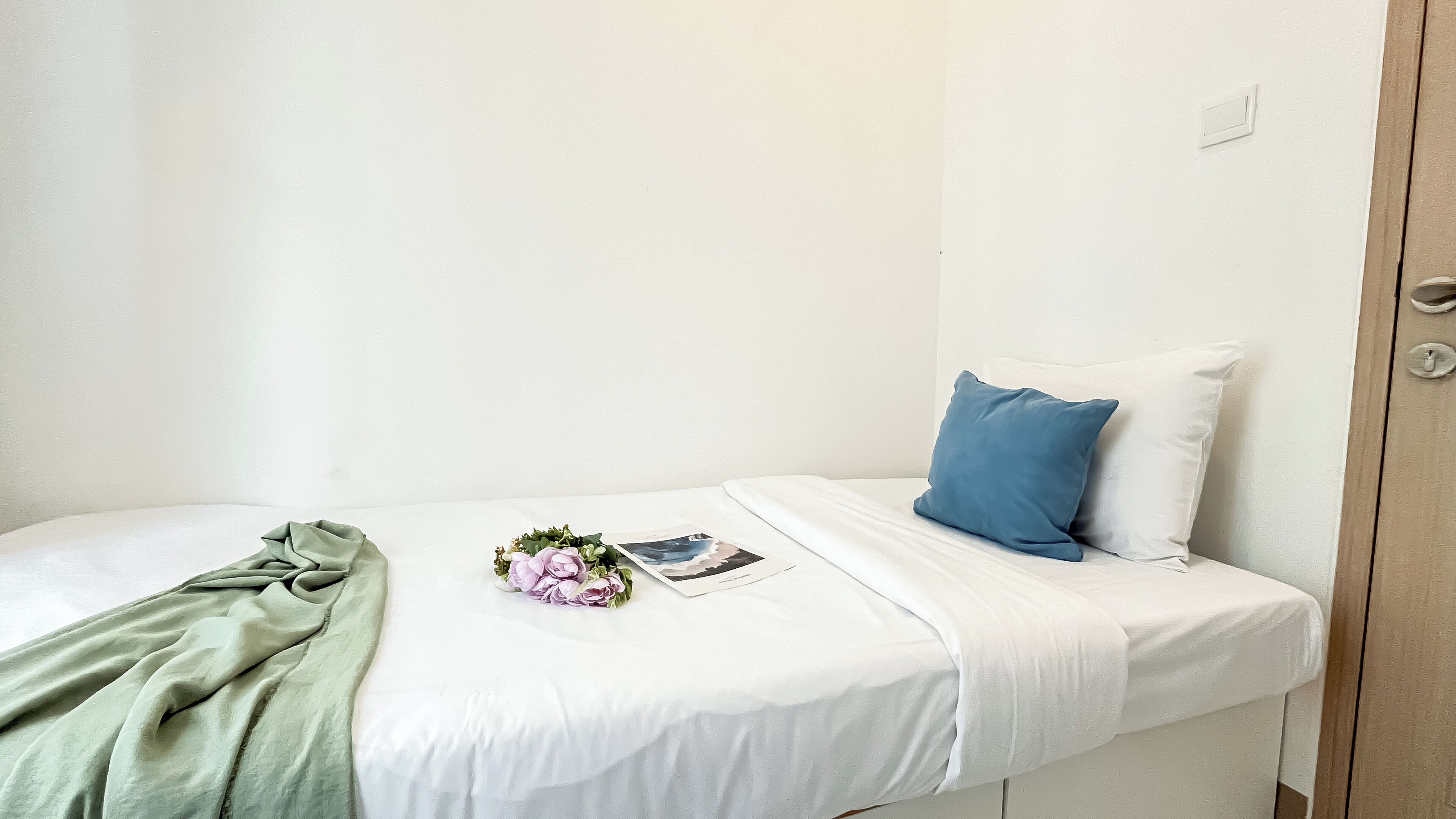 A small, neatly made bed with a white mattress, a blue pillow, and a green blanket. There is a small bouquet of flowers and a magazine on the bed.