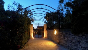 Un sendero de piedra conduce a una entrada porticada con un arco de metal, rodeada de vegetación y muros de piedra.