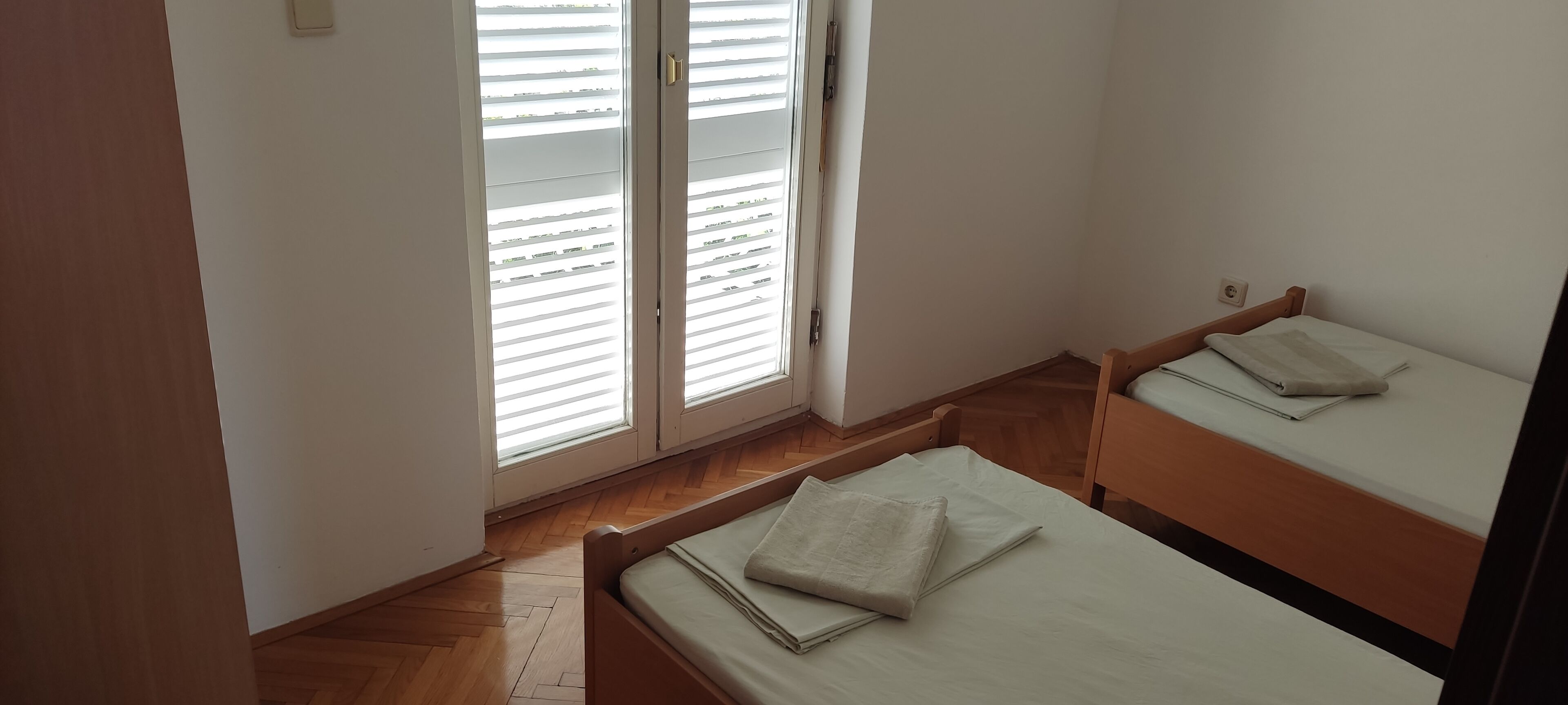 Une petite pièce bien éclairée, avec deux lits simples, un plancher en bois et une grande fenêtre équipée de stores.