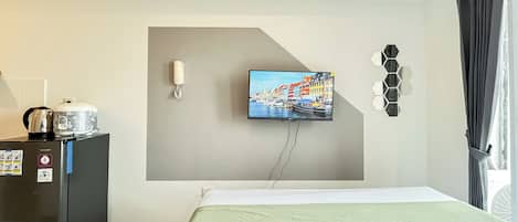 A hotel room with a bed, a TV mounted on the wall, a kettle, a coffee maker, and a small table with a plant.