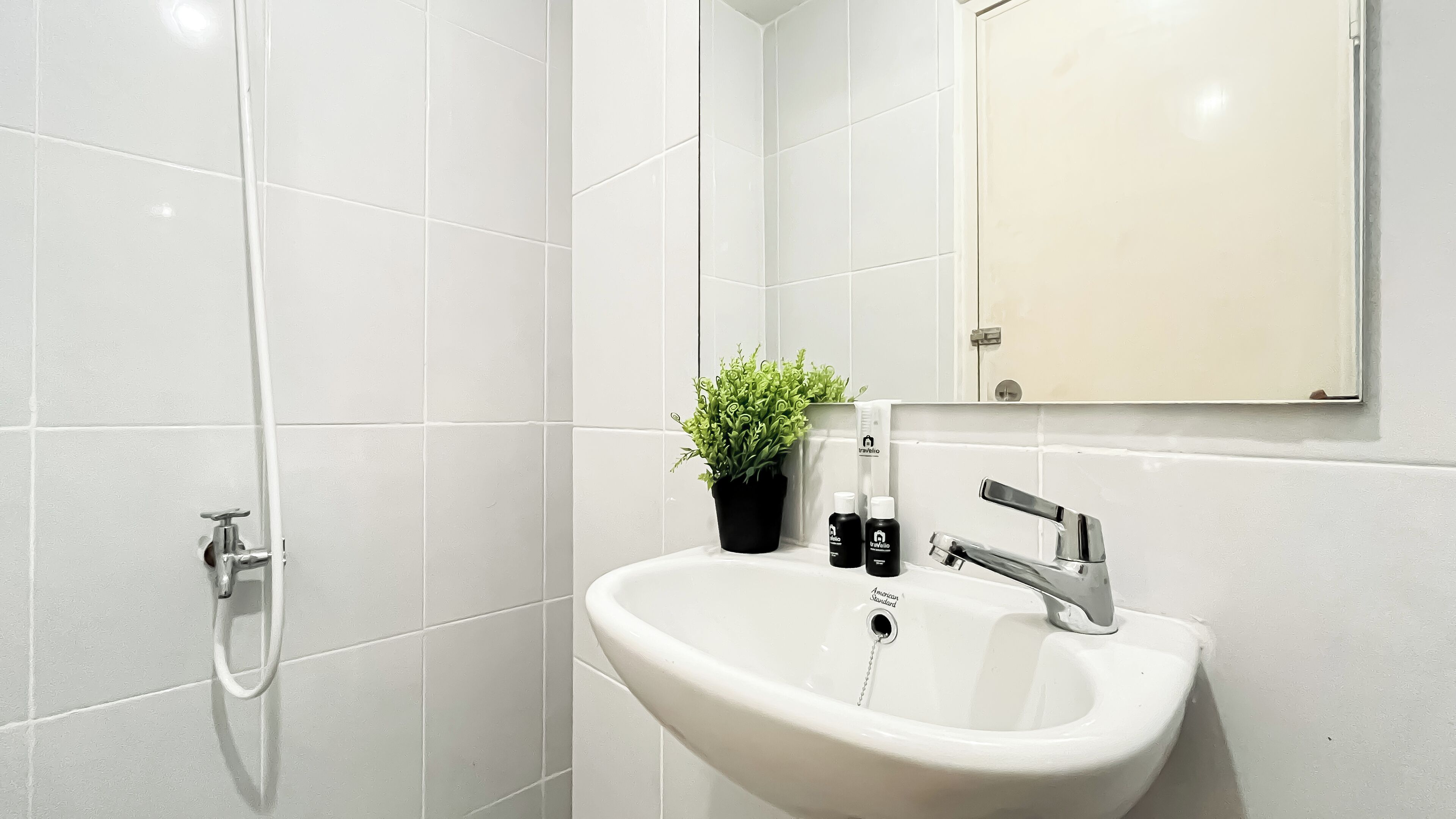 A modern bathroom with white tiles, a sink with a chrome faucet, a potted plant, and toiletries.
