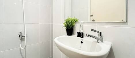 A modern bathroom with white tiles, a sink with a chrome faucet, a potted plant, and toiletries.