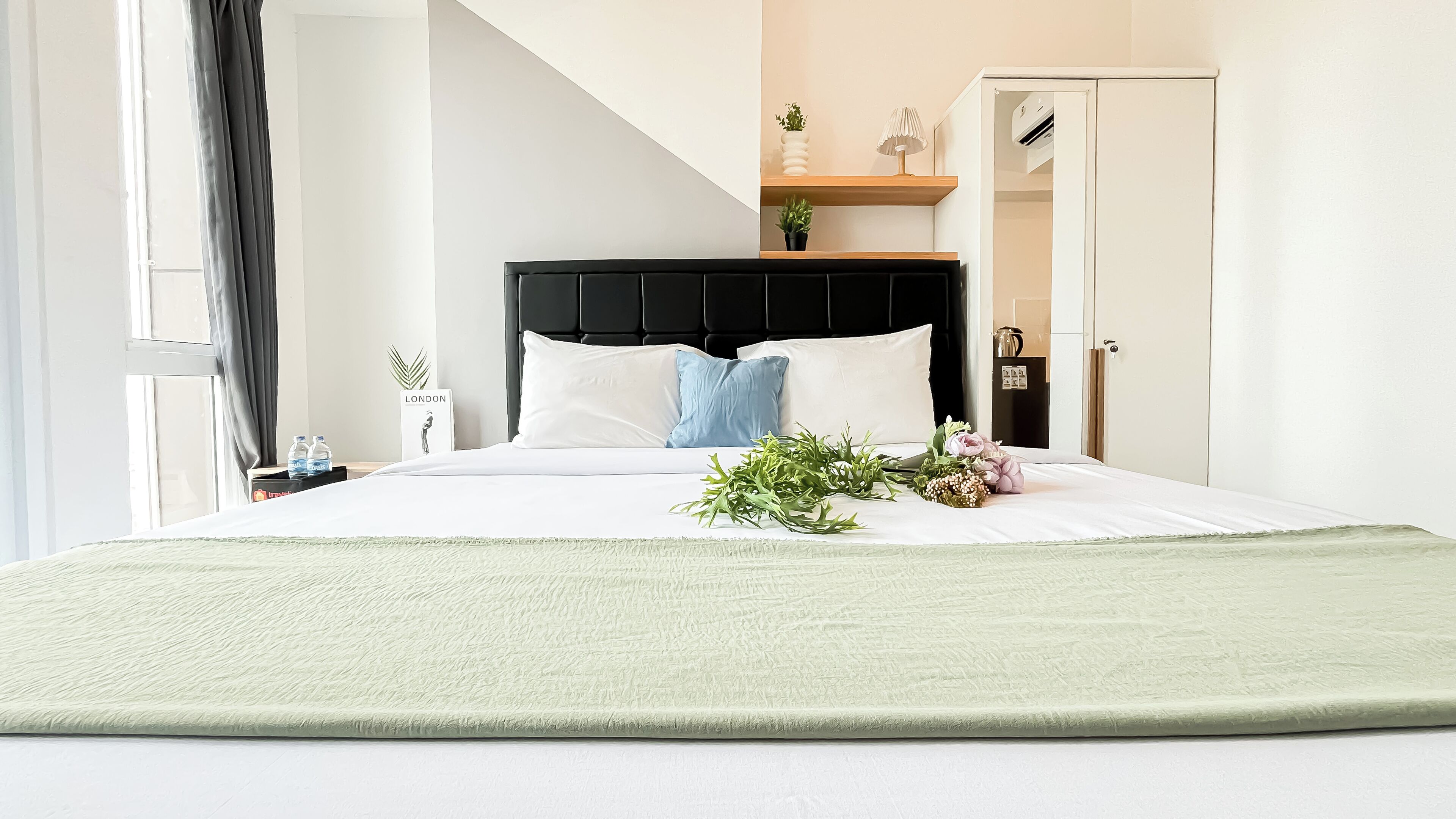 A modern hotel room with a bed, a nightstand, a window with curtains, a door, and a small plant on a shelf.