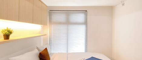 Ein Hotelzimmer mit einem Bett, einem Nachttisch mit Pflanze, einem Fenster mit Jalousien und einer Klimaanlage an der Decke.