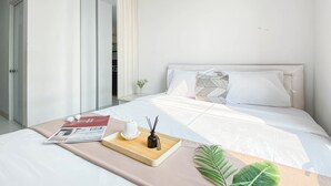A modern hotel room with a neatly made bed, a bedside table with a tray, a magazine, a small vase with flowers, and a potted plant. - Basic Studio Room At Azalea Suites Apartment (Cikarang)