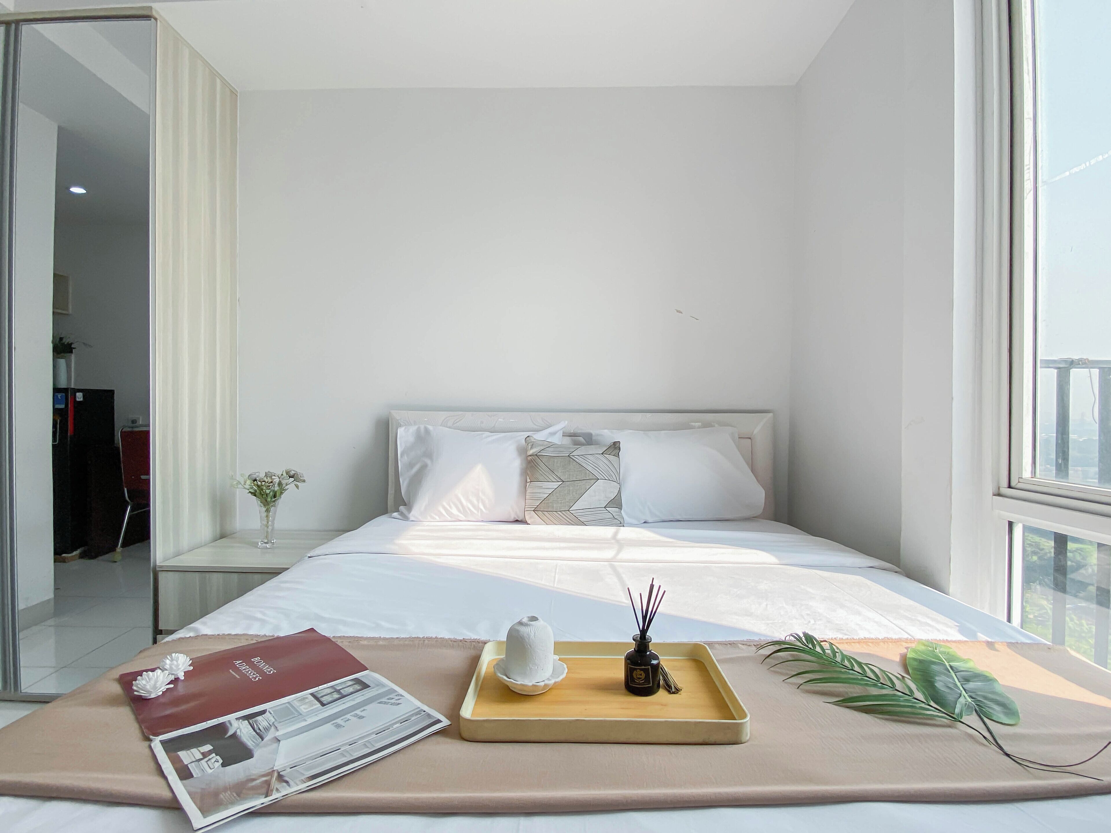 Une chambre d’hôtel moderne avec un lit soigneusement fait, un plateau avec une bouteille et des bâtonnets d’encens, un magazine sur le lit et une plante sur le rebord de la fenêtre.