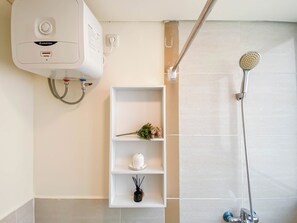 Un baño con un calentador blanco, una ducha con cabezal de ducha manual y un estante con una flor y una vela.
