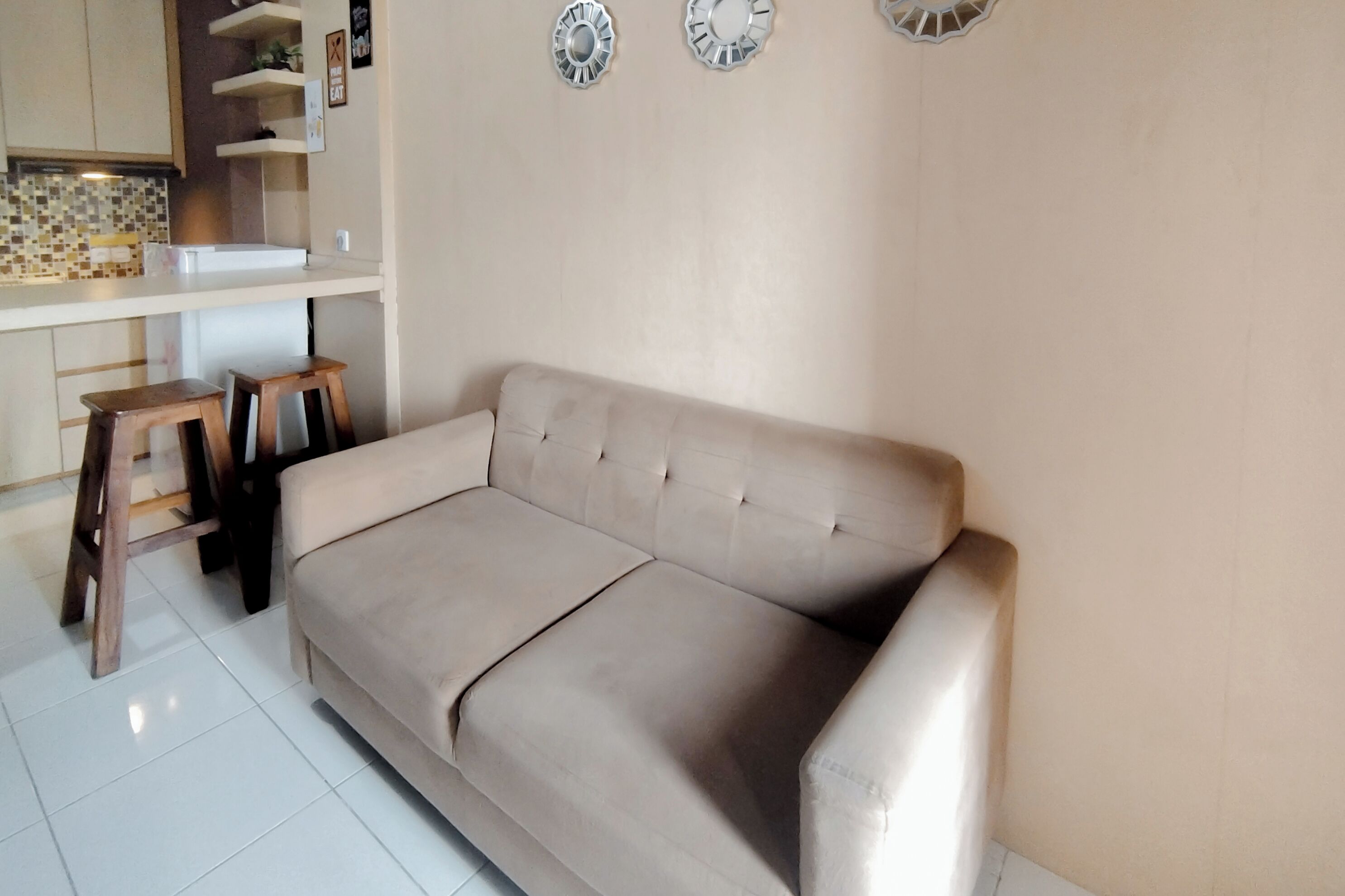 A beige sofa with a cushioned backrest, a wooden stool, and a kitchen area with a tiled backsplash and a white countertop.