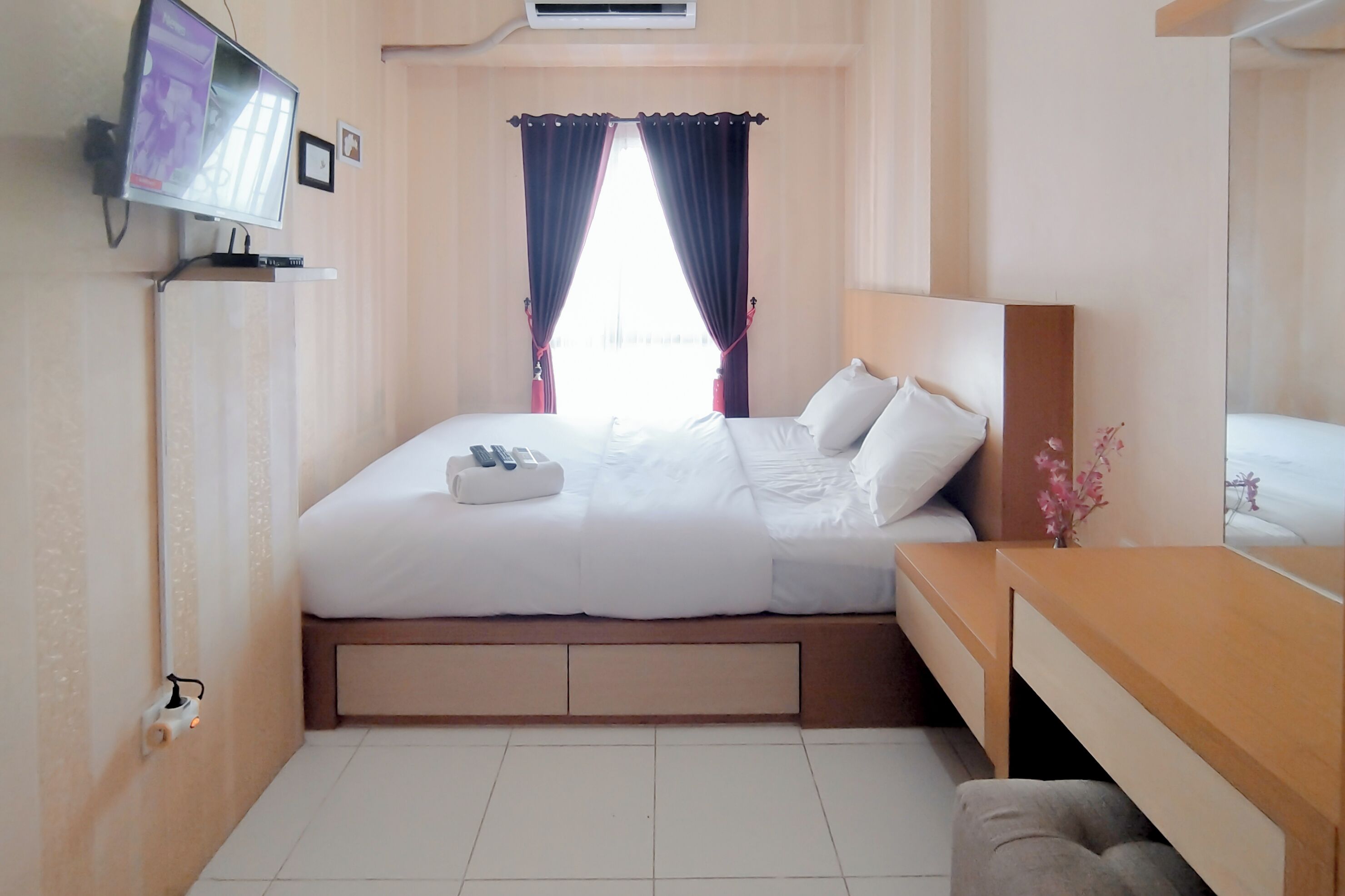 A hotel room with a bed, a TV mounted on the wall, a window with curtains, a bedside table, and a mirror.