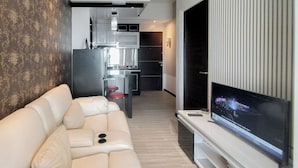 A modern living room with a white leather sofa, a flat-screen TV, and a kitchen in the background. - Monochromatic Modern 1Br At Tamansari Prospero Sidoarjo Apartment (Sidoarjo)