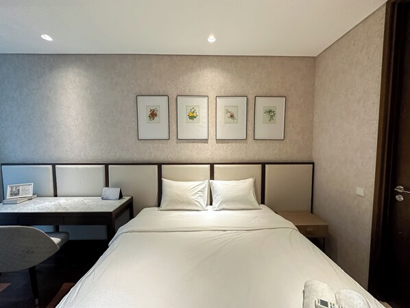 A hotel room with a bed, bedside tables, a desk with a chair, and framed floral artwork on the wall. - Tranquil And Modern Haven 2Br At The Rosebay Apartment (Surabaya)