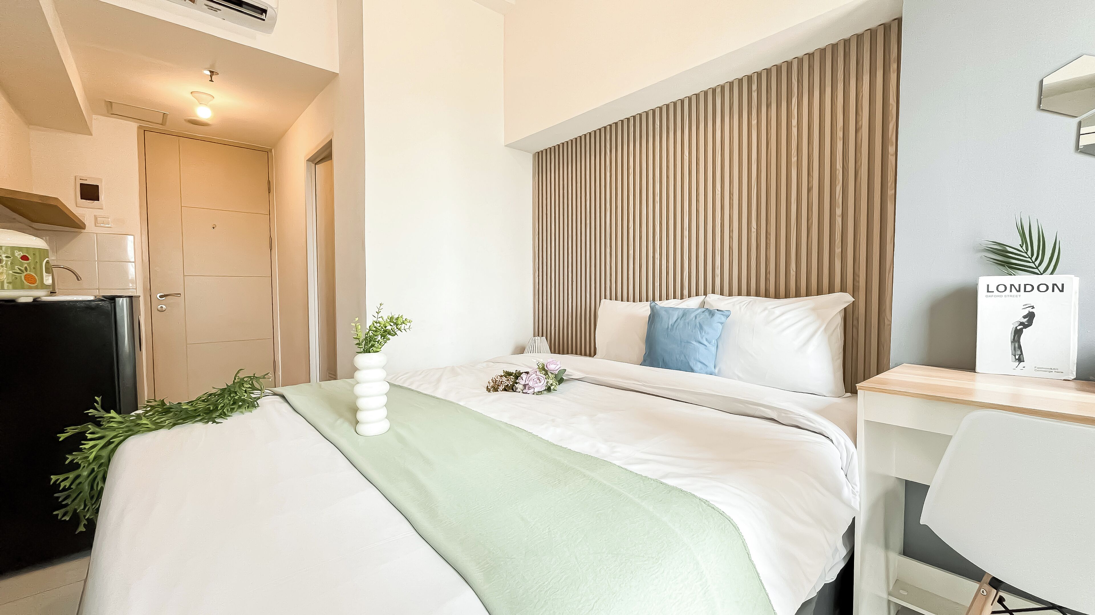 Quarto de hotel com cama, escrivaninha, planta e livro intitulado 'LONDON' sobre a escrivaninha.