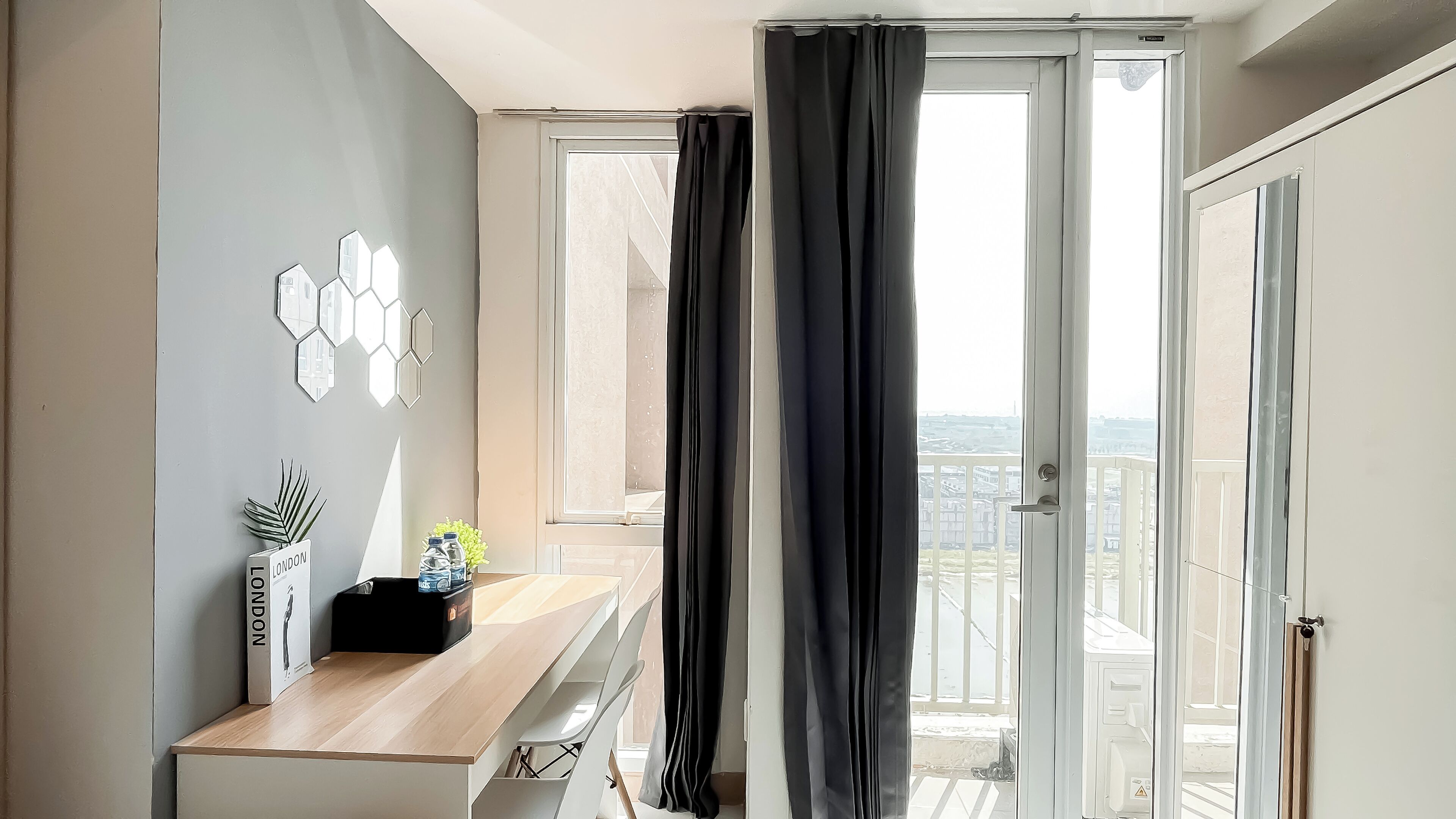 Quarto moderno com escrivaninha de madeira, vista da cidade por grandes janelas e varanda com guarda-corpo.
