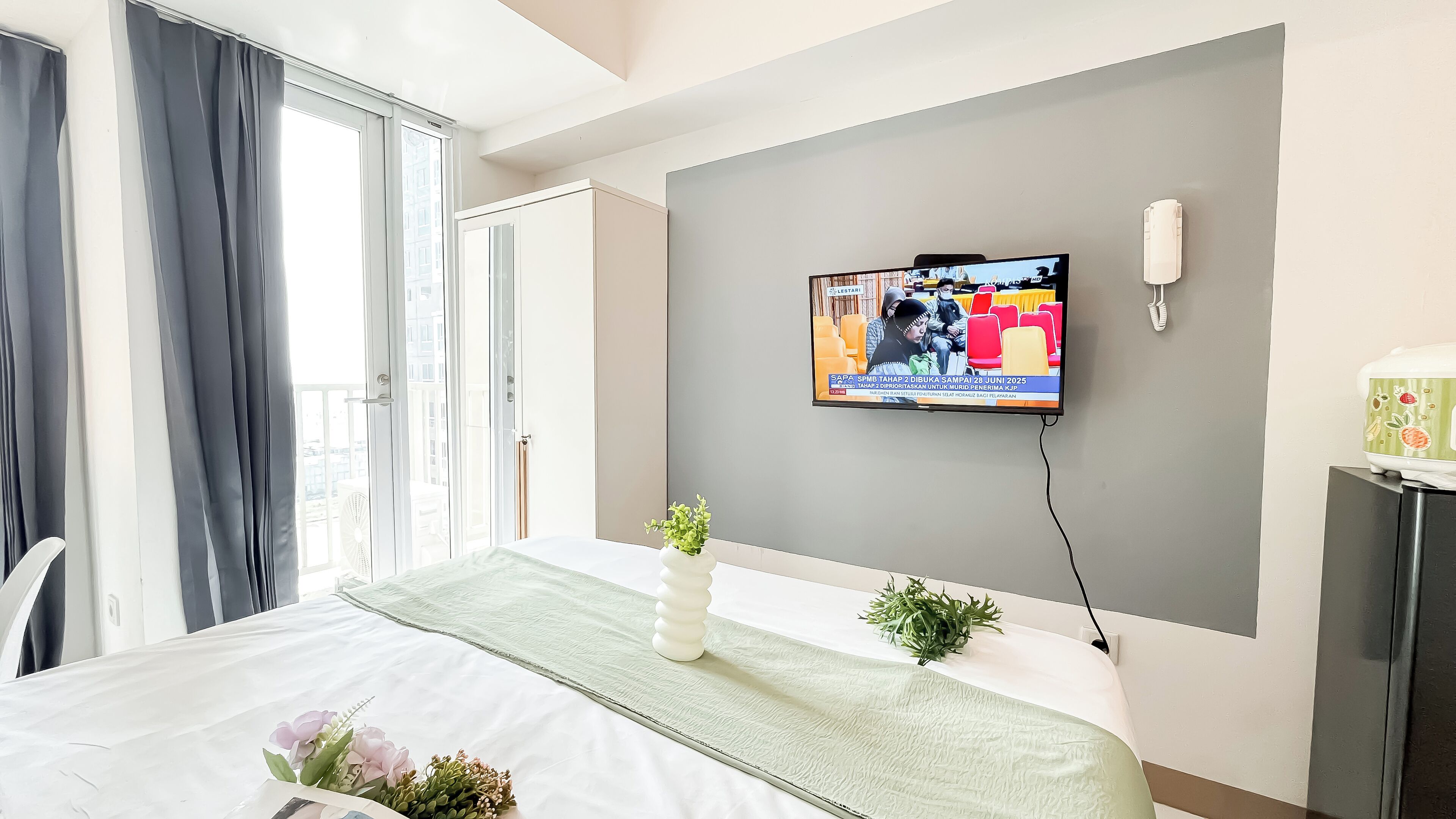 A hotel room with a bed, a TV mounted on the wall, a small plant on the bed, and a window with curtains.