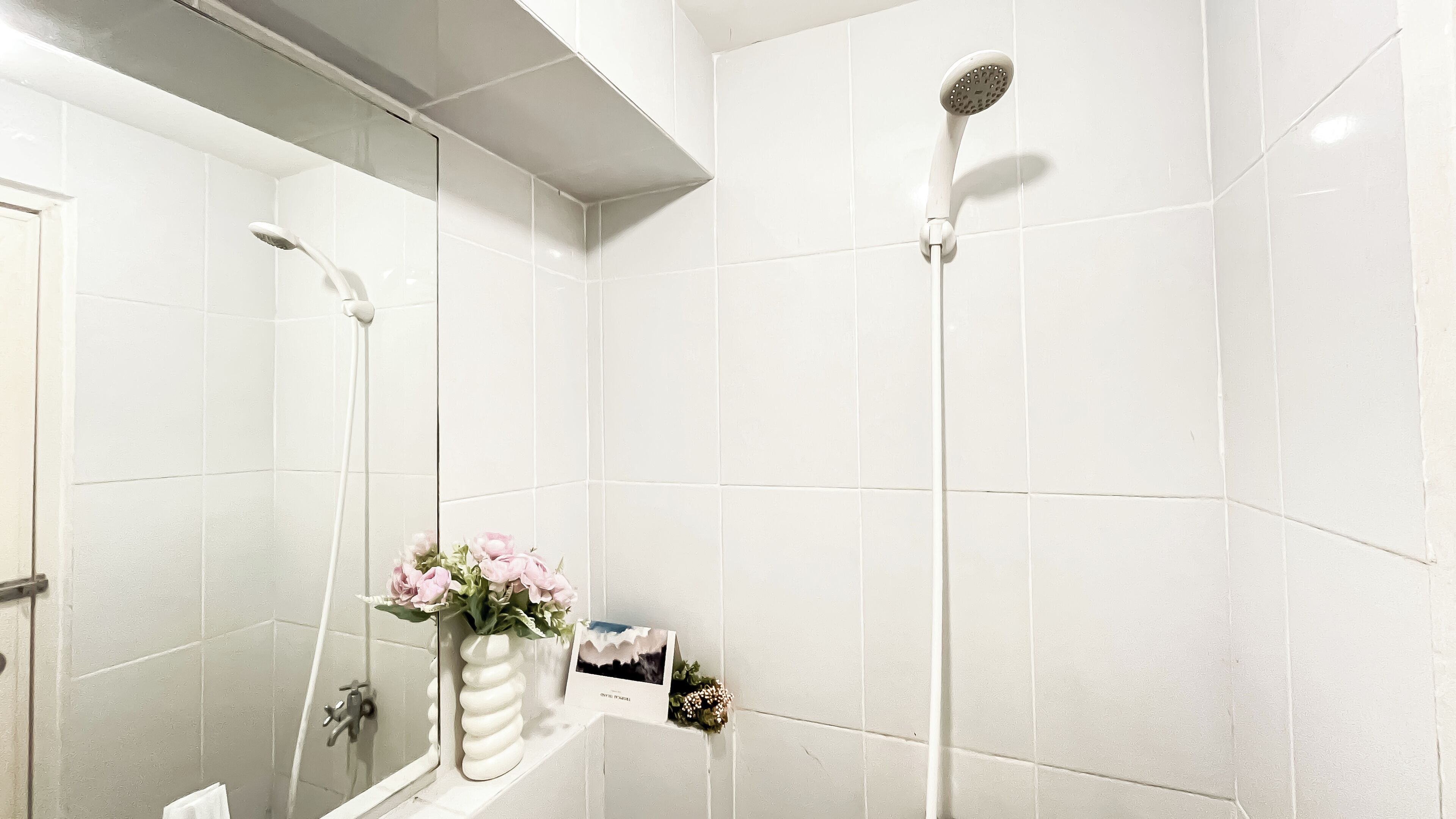 A bathroom with white tiles, a shower, a mirror, and a vase with flowers.