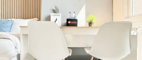A modern hotel room with a bed, two white chairs, a desk with a laptop, a plant, and a travel bag.