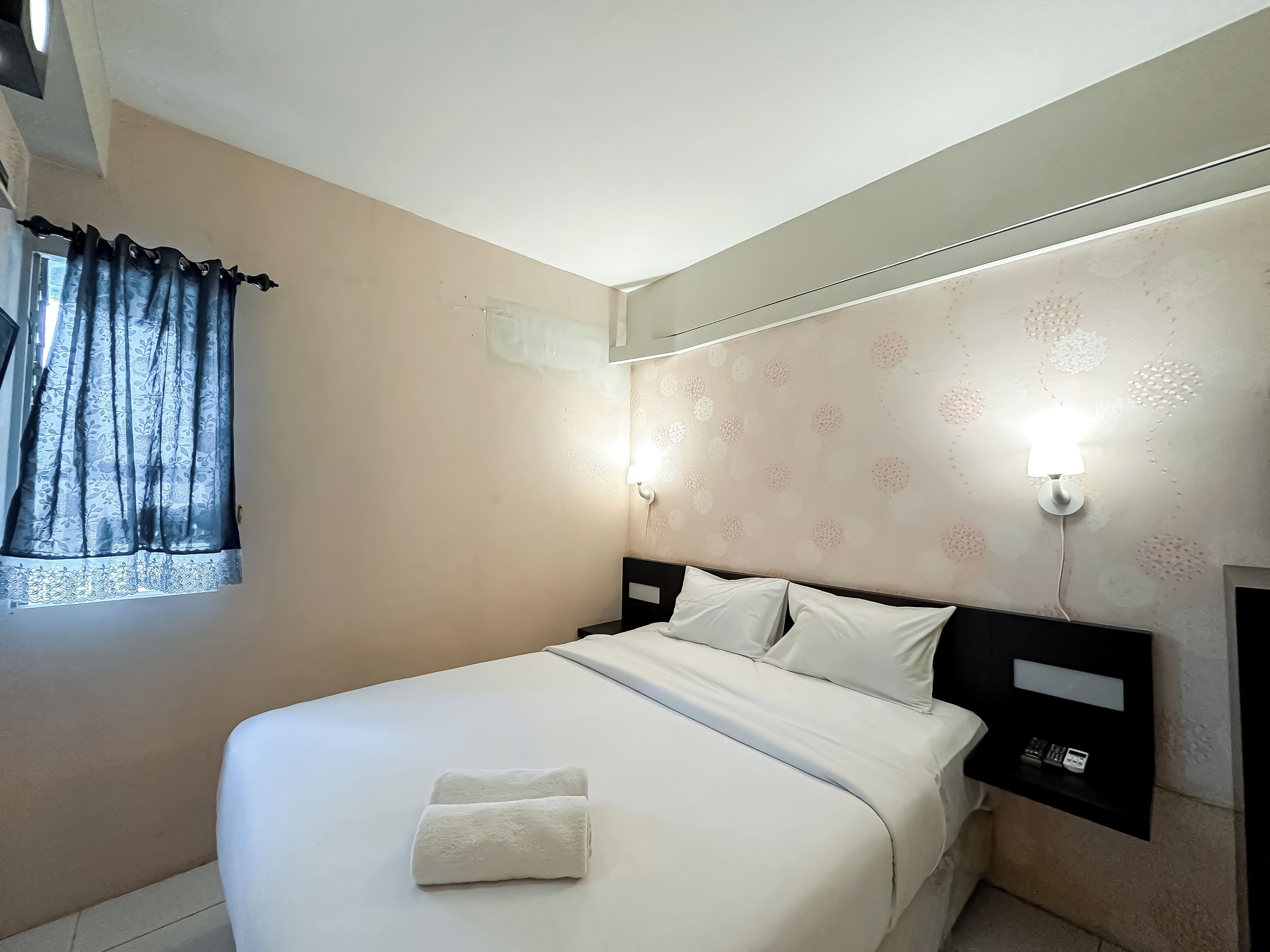 Un lit bien fait, avec du linge de lit blanc et deux serviettes, une table de chevet avec une lampe, et un mur orné d’un motif floral.