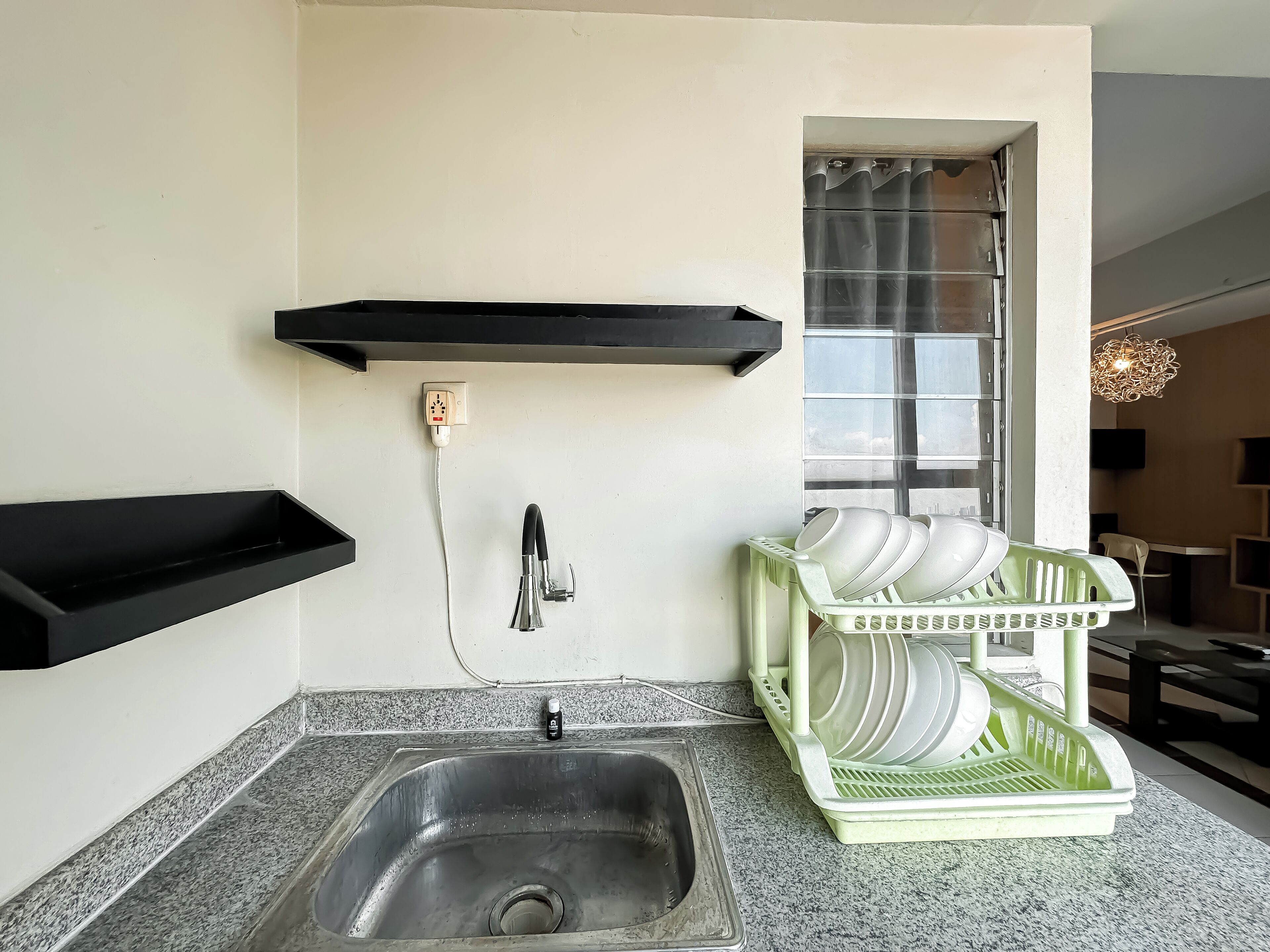 A kitchen with a stainless steel sink, a black shelf, a dish rack, and a window with curtains.