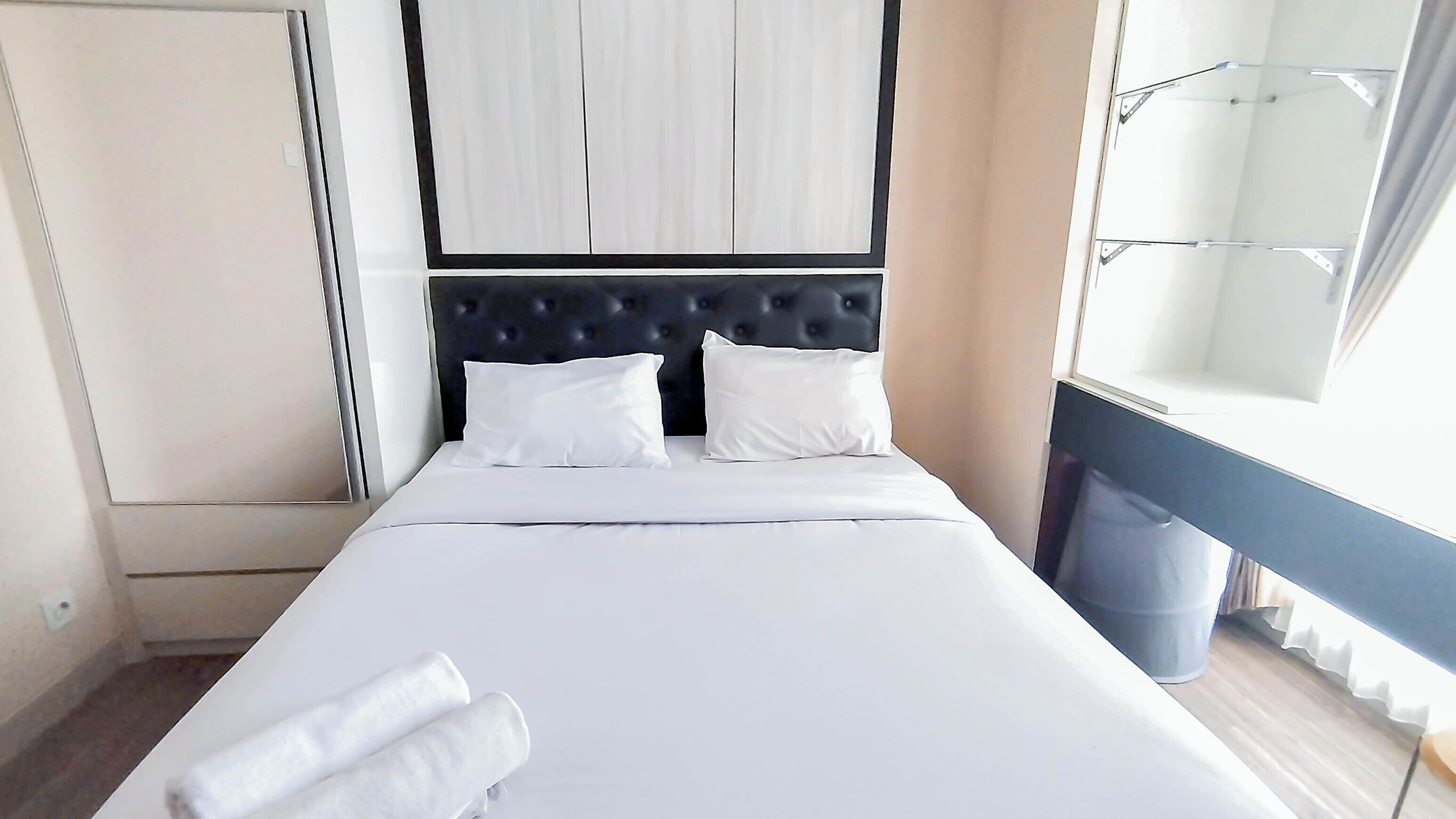 A hotel room with a bed, two pillows, a headboard, a window with curtains, and a trash bin.