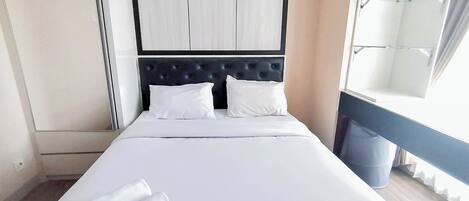 A hotel room with a bed, two pillows, a headboard, a window with curtains, and a trash bin.